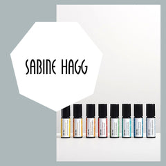 Logo Sabine Hagg und im Hintergrund stehen 8 kleine Flaschem gefüllt mit Naturessenzen in den Farben Gelb, orange dunkel-orange, rot, violett, blau, grün und dunkelblau