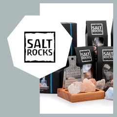 Logo von Saltrocks. Im Hintergrund steht eine Salzreibe aus aus Edelstahl mit einem Holzgefäss aus Eschenholz. Salzbrocken in rosa, blau und braun sind vor Salzpackungen aufgestellt.