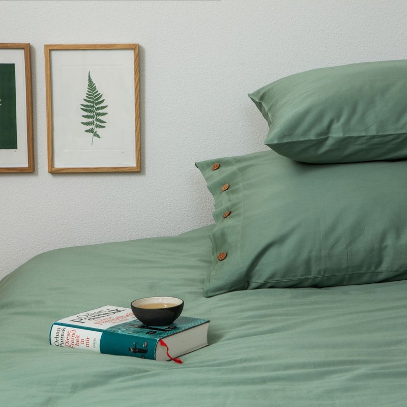 Bettwäsche in Lindengrün mit zwei Kissen, einem Duvet, einem Buch inkl. Teeschale und dahinter 2 Bilder mit Baumsujets